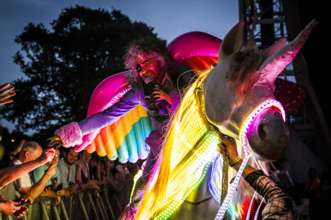 Man singing with rainbow angel wings riding a horse dressed as a unicorn.