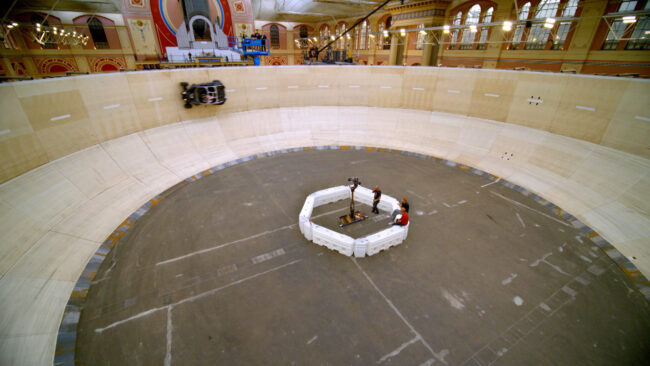 Velodrome with jeep driving around the edge in a stunt whilst a group of people film it.