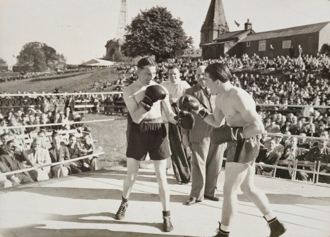 Two boxers fighting in a ring surrounded by an audience watching on a sunny day.