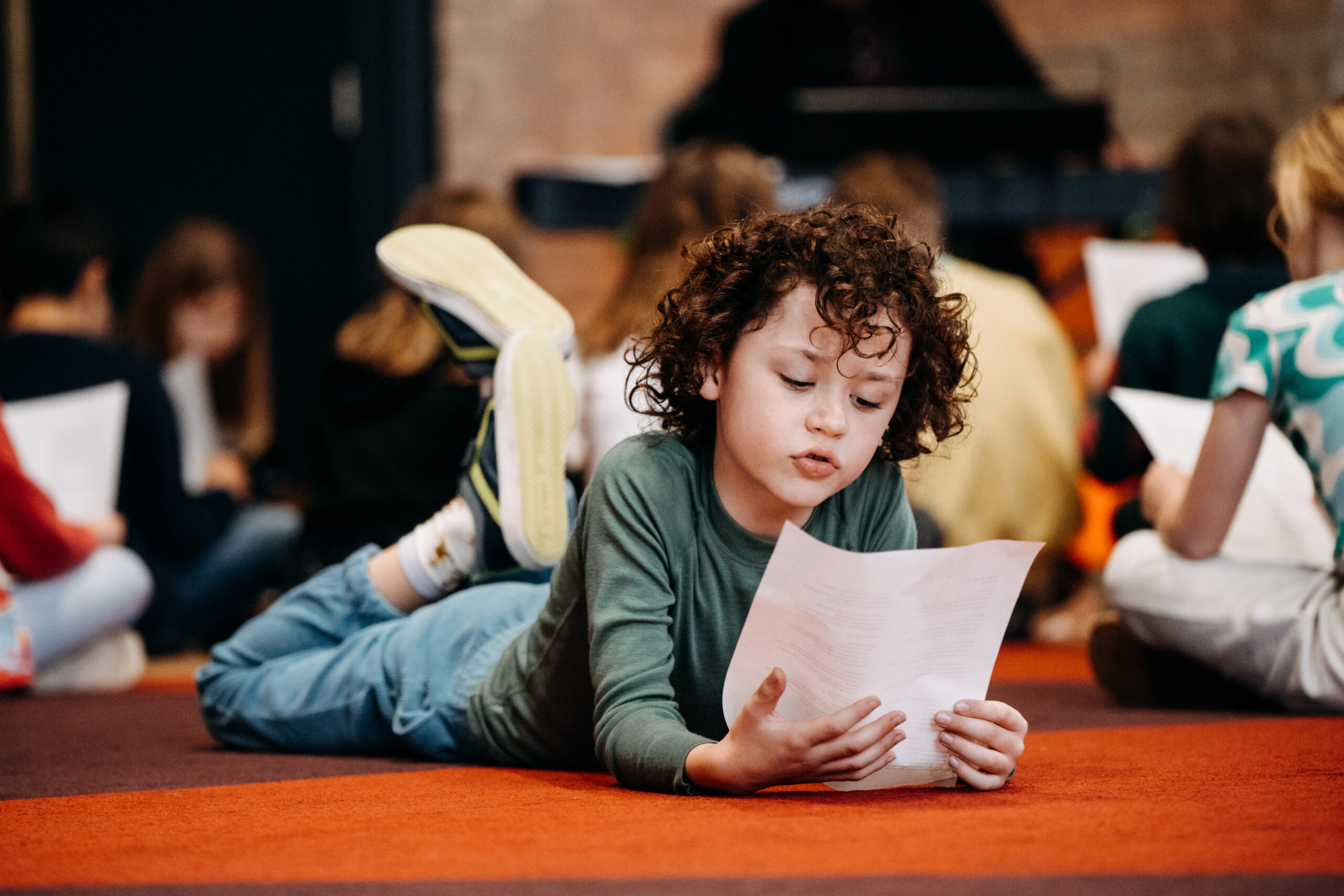 Child with curly hair and grey long sleeved top sits on their stomach looking at a piece of paper