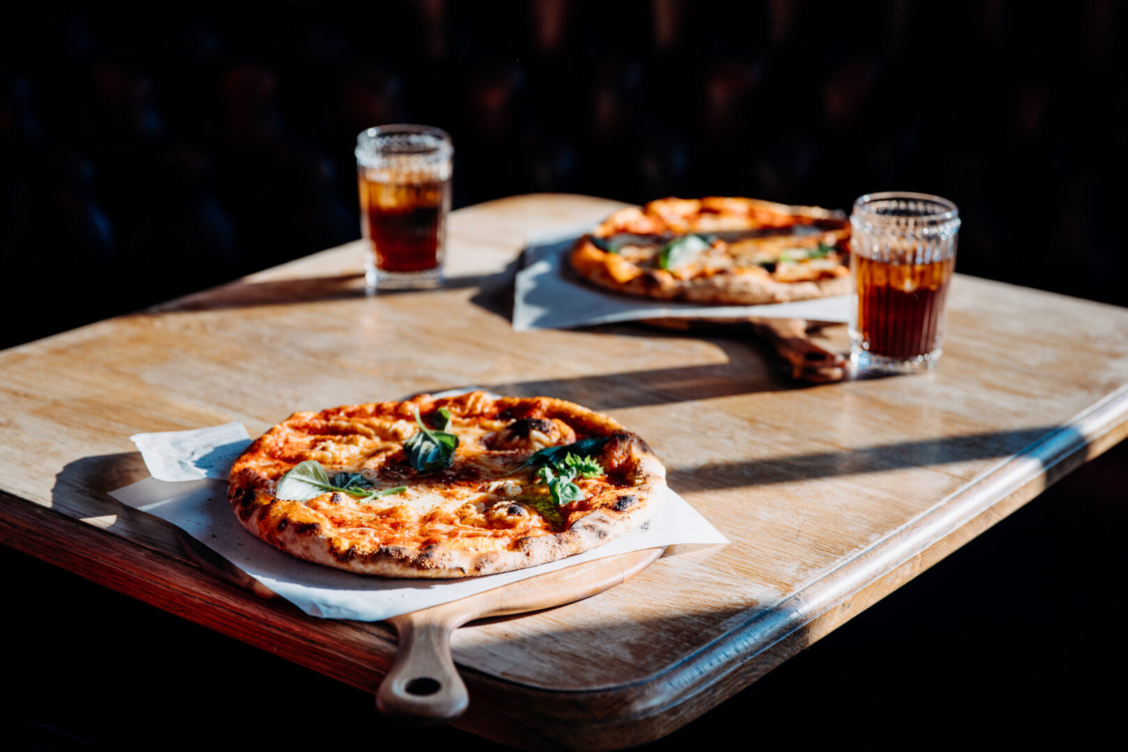 Two pizzas on a table with two glasses of fizzy drink in the sun.