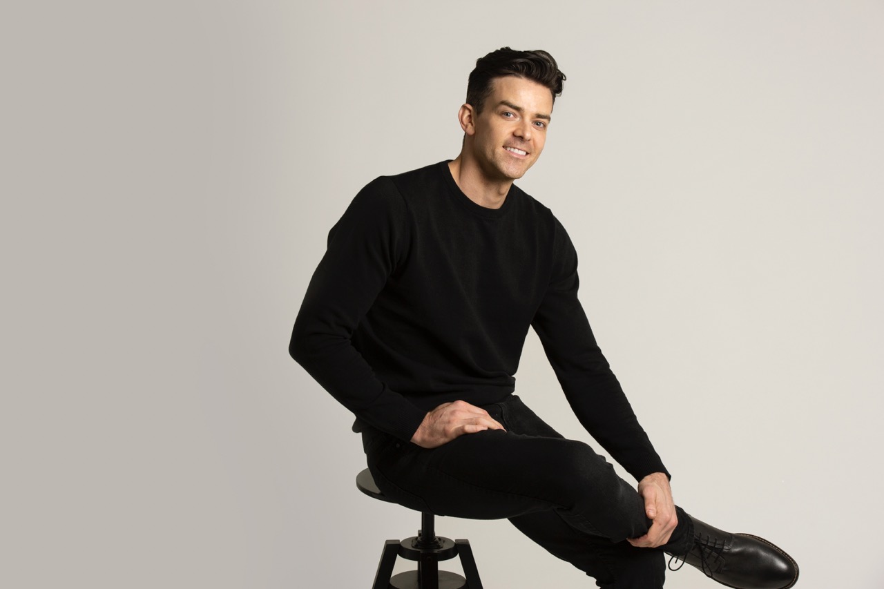 man with dark hair and black top and trousers sits on a stool smiling
