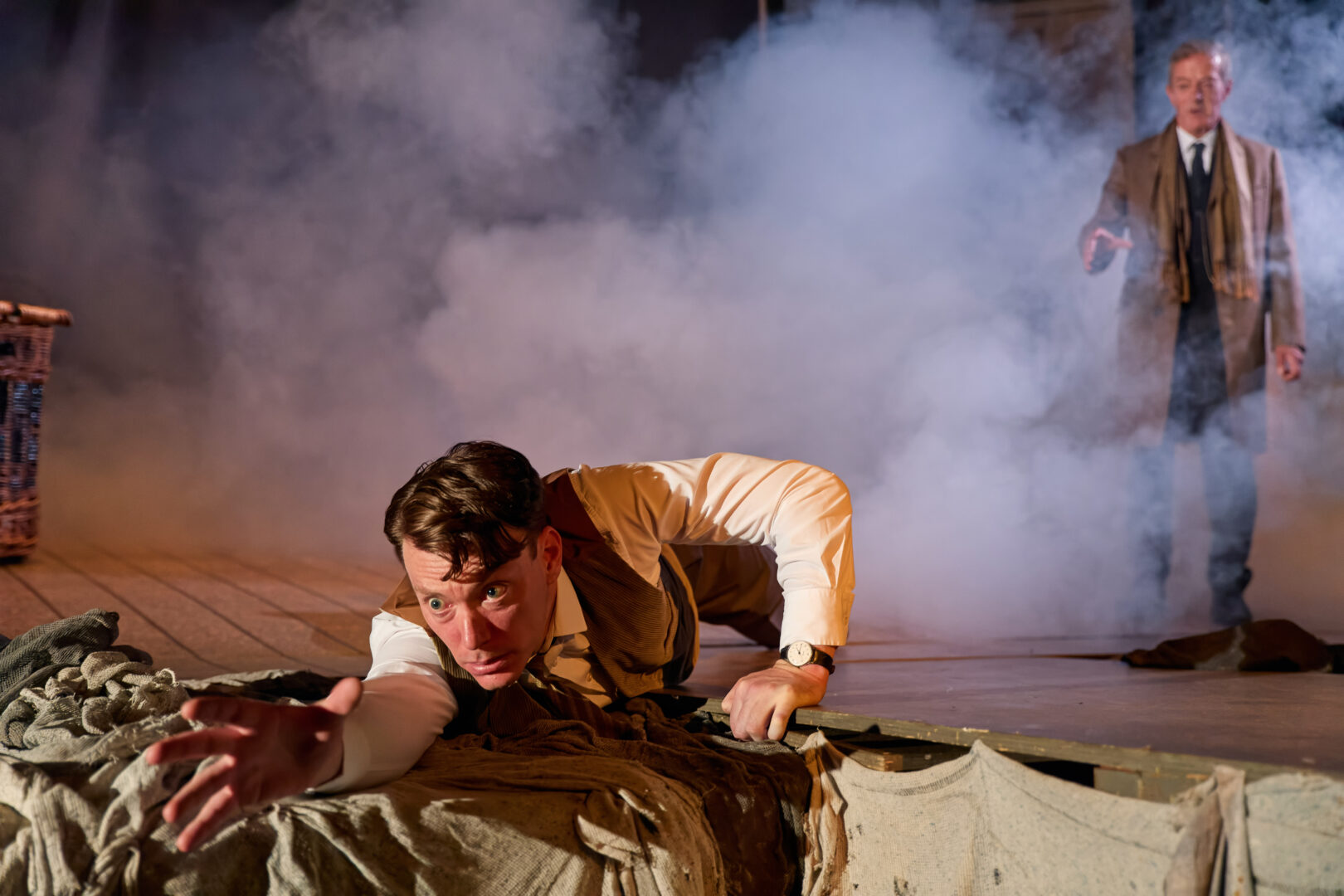 A man crawls forward with a determined expression amidst smoky stage effects, reaching out. A second man stands in the background, also reaching out his hand in uncertainty.
